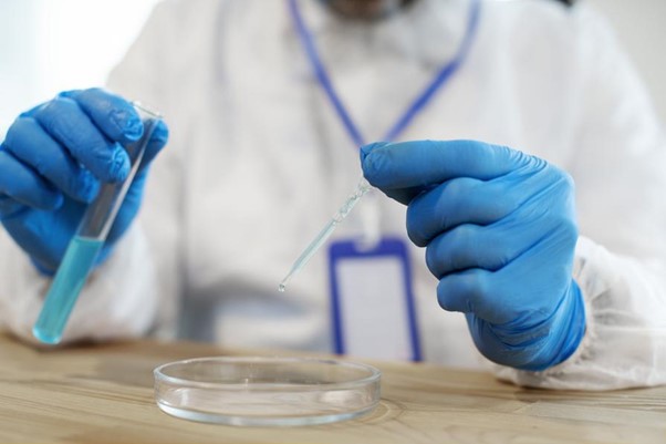 Vedec v ochrannom odeve vykonáva experiment, pri ktorom pipetou kvapká tekutinu z skúmavky do Petriho misky.